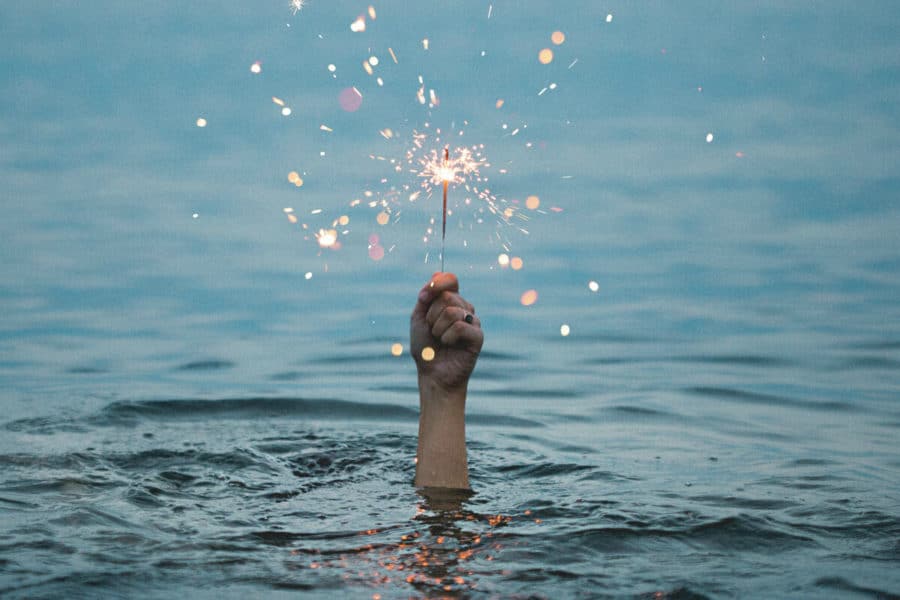 A hand in the centre, holding a lit sparkler above a body of water, signifying "spark new ideas and get a fresh perspective".