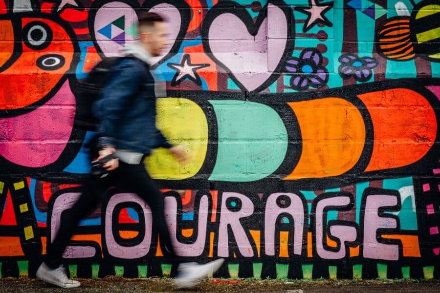 Young white man in jeans and jacket walking left to right past wall covered in colourful street art, with the word 'courage' spray painted in pink at the bottom.