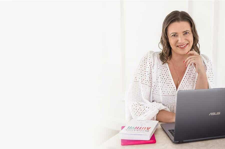 Woman writer wearing white, sitting at a desk with a laptop in front of her.